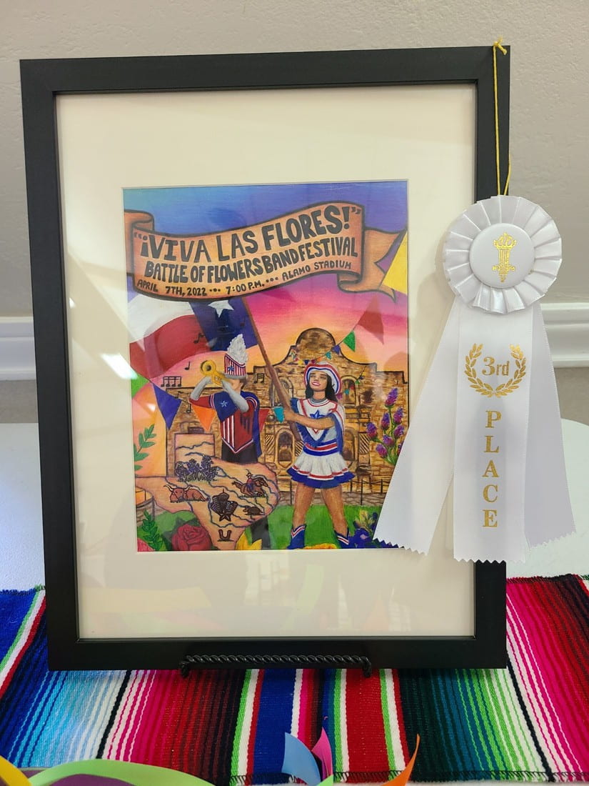 Hailey Hickersons Battle of the Flowers band festival artwork is displayed in a black picture frame on a colorful tablecloth, garnished with a white third place ribbon.